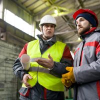 Low angle portrait of two industrial workers wearing warm jackets and hardhats discussing production standing in workshop, copy space
